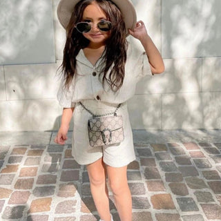Child wearing a white dress, hat, and sunglasses with a small handbag on a stone pavement.